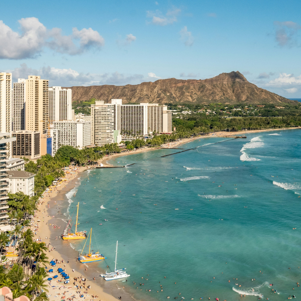 Aerial view of Waikiki Beach in Honolulu with turquoise water, beachfront hotels, palm-lined shoreline, and Diamond Head crater in the background.